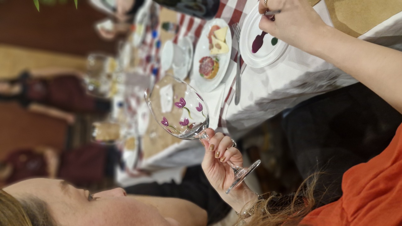 Mãos pintando flores roxas em uma taça de vinho de vidro durante oficina em uma mesa com toalha xadrez